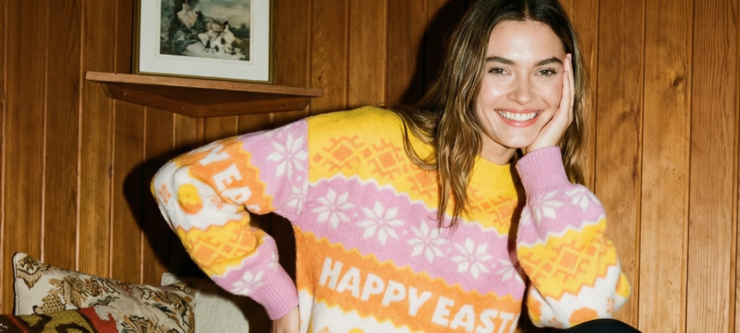 Hymyilevä nainen pukeutuneena vaaleanpunaiseen ja keltaiseen Happy Easter -neuleeseen puupaneeliseinän edessä